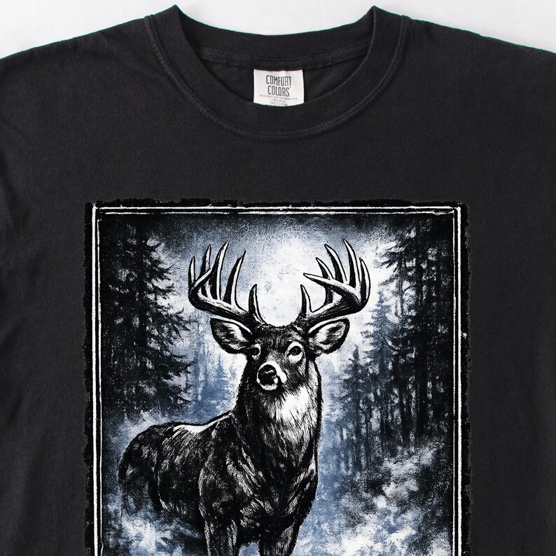 K&ouml;nnte beinhalten: Schwarzes T-Shirt mit einer detaillierten Illustration eines Hirsches mit gro&szlig;em Geweih. Die Grafik ist vor dem Hintergrund einer Waldszene platziert. Das T-Shirt hat einen Rundhalsausschnitt und das Comfort Colors-Logo ist sichtbar.