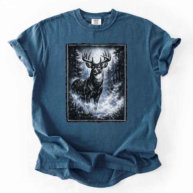 K&ouml;nnte beinhalten: Blaues T-Shirt mit einer Schwarz-Wei&szlig;-Illustration eines Hirsches in einem Wald. Der Hirsch hat ein gro&szlig;es Geweih und steht in einer nebligen Umgebung. Die Grafik ist mit einem schwarzen Rahmen versehen.