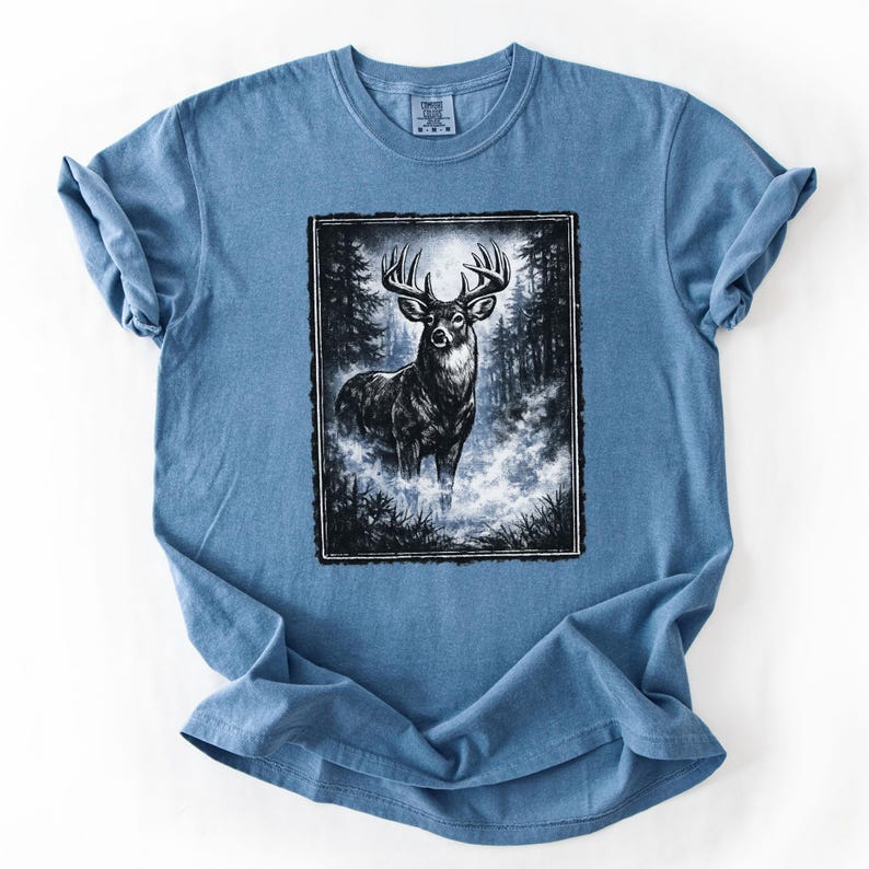 K&ouml;nnte beinhalten: Hellblaues T-Shirt mit einer Schwarz-Wei&szlig;-Illustration eines Hirsches in einer Waldszene. Der Hirsch hat ein gro&szlig;es Geweih und steht in einer nebligen Umgebung. Das T-Shirt hat hochgekrempelte &Auml;rmel.
