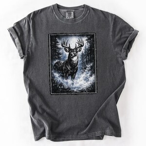 K&ouml;nnte beinhalten: Dunkelgraues T-Shirt mit einer detaillierten Illustration eines Hirsches mit gro&szlig;em Geweih in einer nebligen Waldszene. Die Grafik ist von einem schwarzen Rahmen umgeben, der einen Kontrast zur Farbe des T-Shirts bildet.