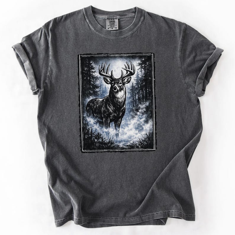 K&ouml;nnte beinhalten: Dunkelgraues T-Shirt mit einer detaillierten Illustration eines Hirsches mit gro&szlig;em Geweih in einer nebligen Waldszene. Die Grafik ist von einem schwarzen Rahmen umgeben, der einen Kontrast zur Farbe des T-Shirts bildet.