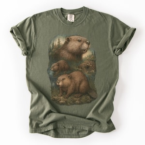 May include: An olive green t-shirt with a detailed illustration of three beavers in a naturalistic setting. The artwork includes a beaver lodge, trees, and water, creating a nature-themed design. The shirt has a classic crew neck and short sleeves.