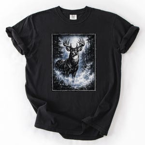 K&ouml;nnte beinhalten: Schwarzes T-Shirt mit einer detaillierten Illustration eines Hirsches mit gro&szlig;em Geweih in einer Waldlandschaft. Das Kunstwerk ist mit einem wei&szlig;en Rahmen versehen und vor einem Hintergrund aus B&auml;umen und nebligen Blaut&ouml;nen platziert. Das Shirt ist aus weichem Material.