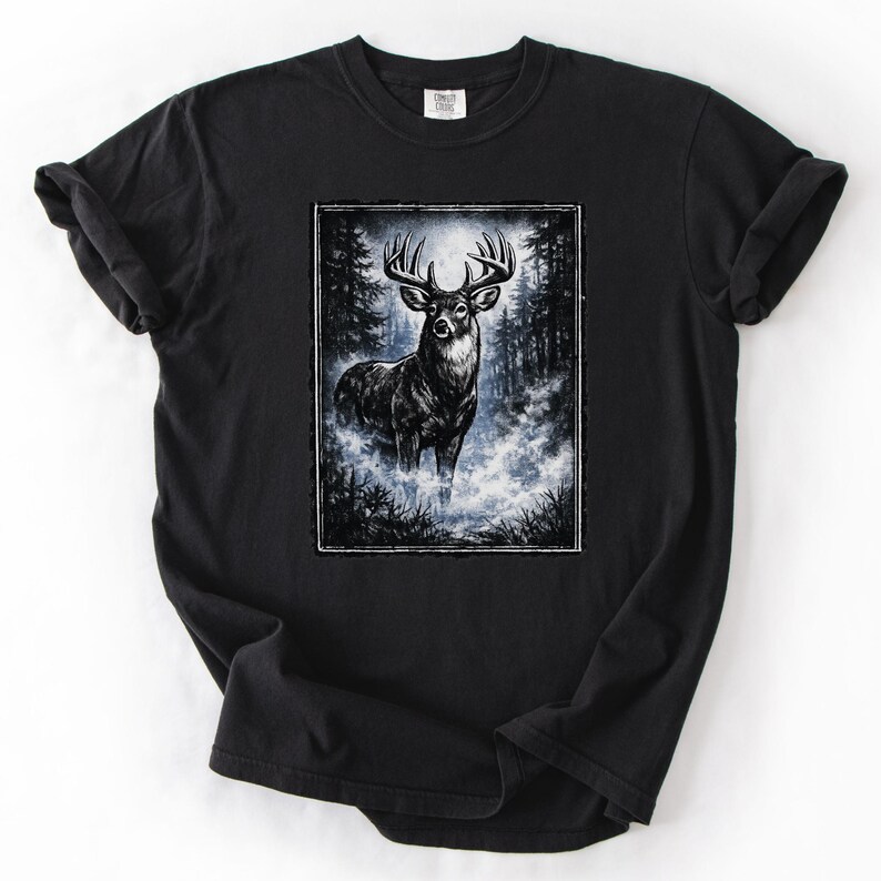 K&ouml;nnte beinhalten: Schwarzes T-Shirt mit einer detaillierten Illustration eines Hirsches mit gro&szlig;em Geweih in einer Waldlandschaft. Das Kunstwerk ist mit einem wei&szlig;en Rahmen versehen und vor einem Hintergrund aus B&auml;umen und nebligen Blaut&ouml;nen platziert. Das Shirt ist aus weichem Material.