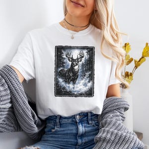 K&ouml;nnte beinhalten: Wei&szlig;es T-Shirt mit einer Schwarz-Wei&szlig;-Illustration eines Hirsches mit gro&szlig;em Geweih in einer Waldszene. Der Hirsch steht im Mittelpunkt, umgeben von B&auml;umen und einer nebligen Atmosph&auml;re. Das T-Shirt ist aus weichem Material.