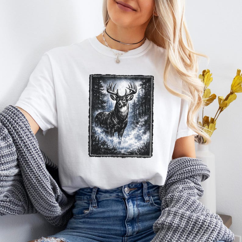 K&ouml;nnte beinhalten: Wei&szlig;es T-Shirt mit einer Schwarz-Wei&szlig;-Illustration eines Hirsches mit gro&szlig;em Geweih in einer Waldszene. Der Hirsch steht im Mittelpunkt, umgeben von B&auml;umen und einer nebligen Atmosph&auml;re. Das T-Shirt ist aus weichem Material.