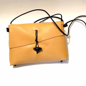 May include: A small, tan leather crossbody bag with a black cord strap. The bag has a flap closure secured by a black toggle. The edges are stitched with a zig-zag pattern. The bag is rectangular and appears to be handmade.