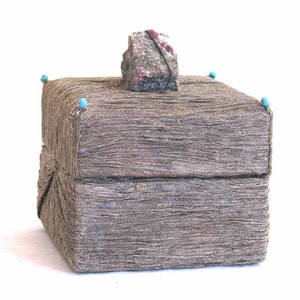 May include: A square, textured box with a lid, covered in a woven, metallic fabric. The lid is topped with a rough, gray stone with red accents and secured with a black string. Small turquoise beads adorn the corners.