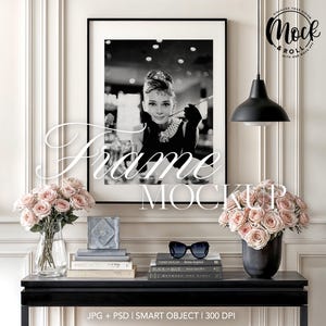 May include: A black and white photo of a woman in a black dress and pearls is framed in a black frame and hanging on a white wall above a black console table. The table has a vase of pink roses, a stack of books, and a pair of sunglasses on it.