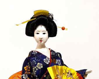 May include: A decorative doll with a pale face and black hair styled in an updo with gold and orange hair ornaments. The doll wears a dark blue kimono featuring a white and gold floral pattern, an orange obi, and holds a gold folding fan.
