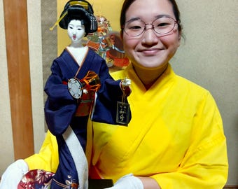 A beautiful and cute geisha doll. She is wearing a purple kimono with afloral pattern anda red obi sash.The traditional kimono size is 47cm.