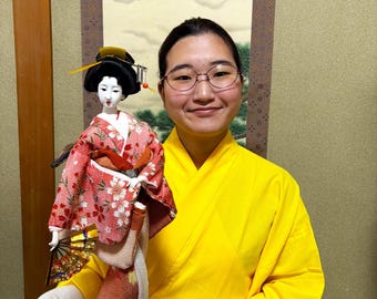 Beautiful and cute geisha doll. Holds a small fan in her hand and is dressed in a luxurious traditional pink costume. Height: 44.5 cm.