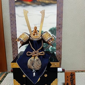 Puede incluir: Un casco de samurái japonés tradicional, o kabuto, con detalles dorados y plateados, expuesto en un soporte de madera negro con adornos dorados. El casco está adornado con un paño azul y una pintura de montaña roja y dorada está en el fondo.