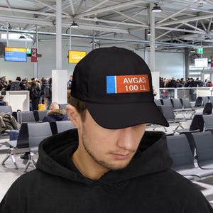Puede incluir: Gorra de béisbol negra con un parche rectangular con el texto "AVGAS 100 LL". El parche tiene una franja vertical azul a la izquierda, una franja naranja en el medio y texto blanco.