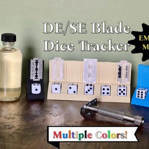 May include: Three different colored dice trackers made from plastic with embedded magnets. Each tracker is shaped like a razor blade holder and has a slot for a razor blade. The trackers are in black, beige, and blue. The text "EMBEDDED MAGNETS" is on a black starburst shape. The text "DE/SE Blade Dice Tracker" is in white letters on a black background. The text "Multiple Colors!" is in rainbow letters on a black background.