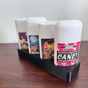 May include: Four white deodorant sticks with colorful labels. The labels feature illustrations and text. The text on the labels reads "Shave", "FRE", "Phoenix Shaving", and "Northpole Barbershop Cane! Artisan Deodorant".