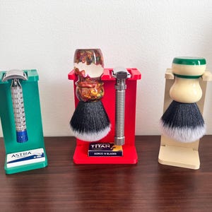May include: Three shaving brush stands, each with a different design. The stand on the left is green and holds a silver razor. The stand in the middle is red and holds a brush with a clear handle and a black and white brush head. The stand on the right is beige and holds a brush with a white handle and a black and white brush head. The stand in the middle also has a pack of razor blades that says "TITAN DORCO 10 BLADES".