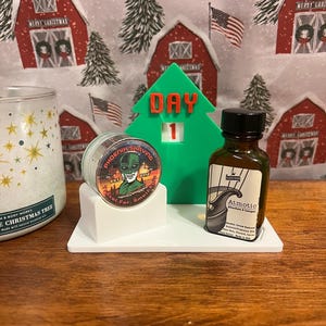 May include: A green Christmas tree-shaped countdown calendar with the number 1 displayed. The calendar is on a white platform with a small jar of shaving soap and a bottle of aftershave. The shaving soap jar has a green alien logo with the text "Phoenix Shaving" and "Est. 2012". The aftershave bottle is brown glass with a white label that says "Atmotic Aftershave & Cologne".