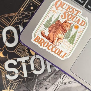 Könnte beinhalten: Ein Aufkleber auf einem Laptop mit einer Cartoon-orangefarbenen Tabby-Katze mit Rucksack, den Worten "Quest Squad Broccoli" sowie Sonne und Bäumen. Der Aufkleber befindet sich auf einem silbernen Laptop neben einem Buch mit Gold- und Weißtext.