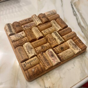 May include: Square cork trivet made from recycled wine corks. The corks are arranged in a mosaic pattern and feature various winery names and logos. The trivet is a natural brown colour and has a cork border.