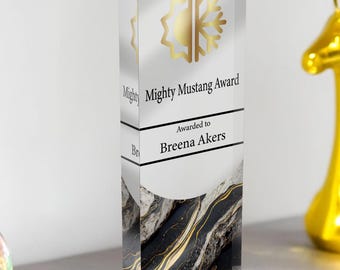May include: A clear acrylic award with a gold and black marble design. The award has the text "Mighty Mustang Award" and "Awarded to Breena Akers". A gold giraffe statue is in the background.