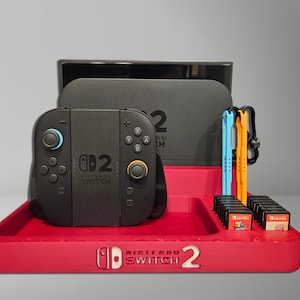Pode incluir: Um organizador vermelho Nintendo Switch 2 contém um console preto, uma capa de transporte preta, cartuchos de jogos e controladores Joy-Con azuis e laranja. O organizador tem o logotipo Nintendo Switch 2.