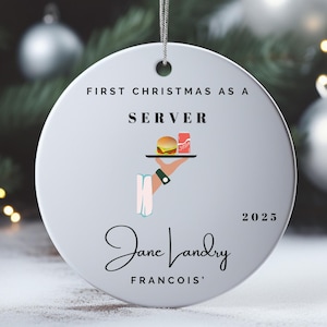 May include: A round, white Christmas ornament with the text "First Christmas as a Server" and a graphic of a burger and soda on a tray. The ornament also includes the name "Jane Landry" and the year "2025".