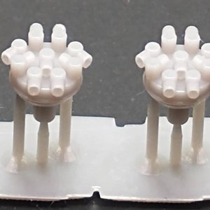 Könnte beinhalten: Zwei weiße Plastik-Modell-Motorteile mit mehreren Zylindern und einem Sockel mit drei Beinen.