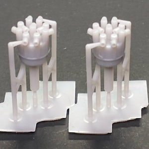 Könnte beinhalten: Zwei weiße Plastikmodelle von Motorblöcken mit mehreren Zylindern. Jeder Block ist auf einem weißen Plastiksockel montiert.