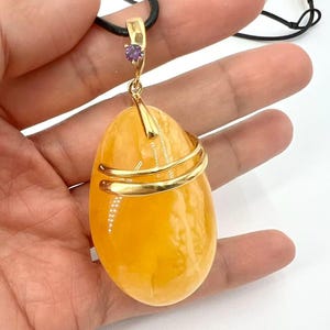 May include: A large, oval amber pendant with a gold-colored metal wrap and a small purple gemstone. The pendant hangs from a black cord necklace. The amber is a translucent, warm yellow-orange color.