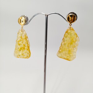 May include: A pair of gold-toned earrings with irregular, textured, yellow-toned pendants. The earrings are displayed on a silver metal stand against a white background. The earrings have a post-back closure.
