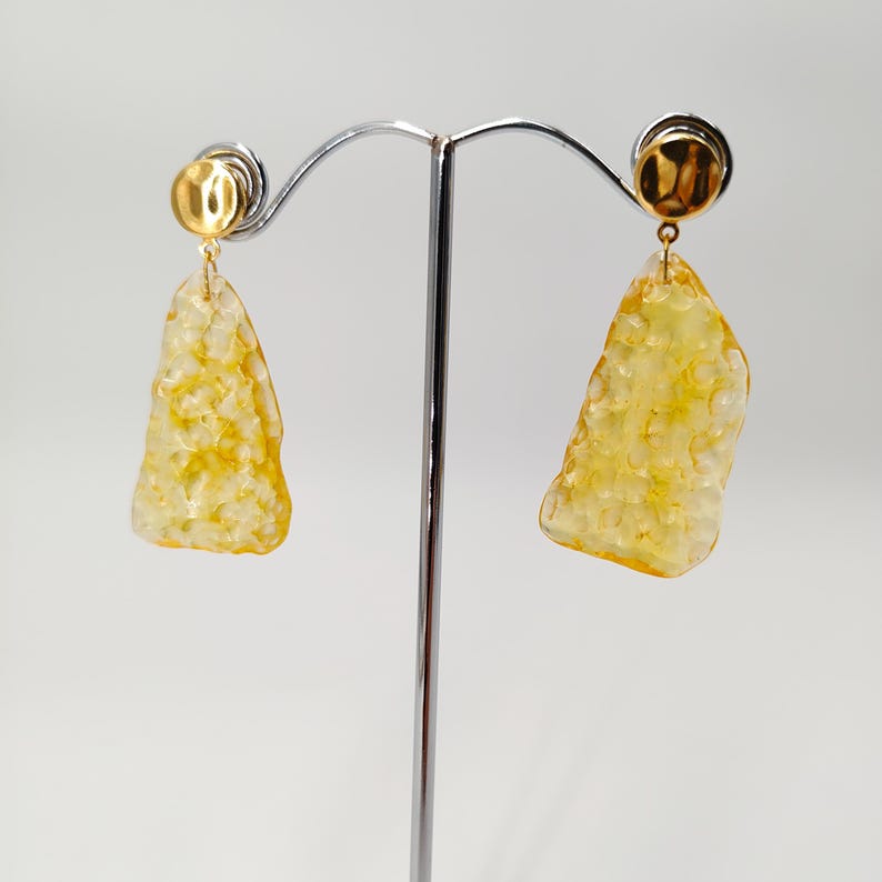 May include: A pair of gold-toned earrings with irregular, textured, yellow-toned pendants. The earrings are displayed on a silver metal stand against a white background. The earrings have a post-back closure.