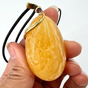May include: A large, oval-shaped pendant with a marbled, amber-colored stone. The pendant is suspended from a black cord and features a gold-colored metal accent with a small purple gemstone. The pendant is held in a hand.