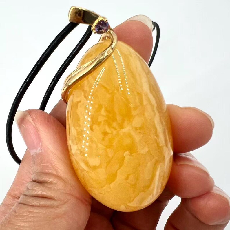 May include: A large, oval-shaped pendant with a marbled, amber-colored stone. The pendant is suspended from a black cord and features a gold-colored metal accent with a small purple gemstone. The pendant is held in a hand.
