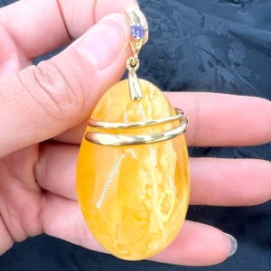 May include: A golden pendant with a large, oval amber stone. The amber is a warm yellow color with natural patterns. A gold band wraps around the stone, and a small purple gemstone is set in the gold bail. The pendant is held by a hand.