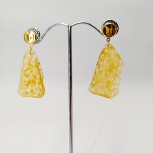 May include: A pair of gold-colored earrings with a textured, irregular, yellow-orange pendant. The earrings are displayed on a silver stand against a white background. The earrings have a post-back closure.