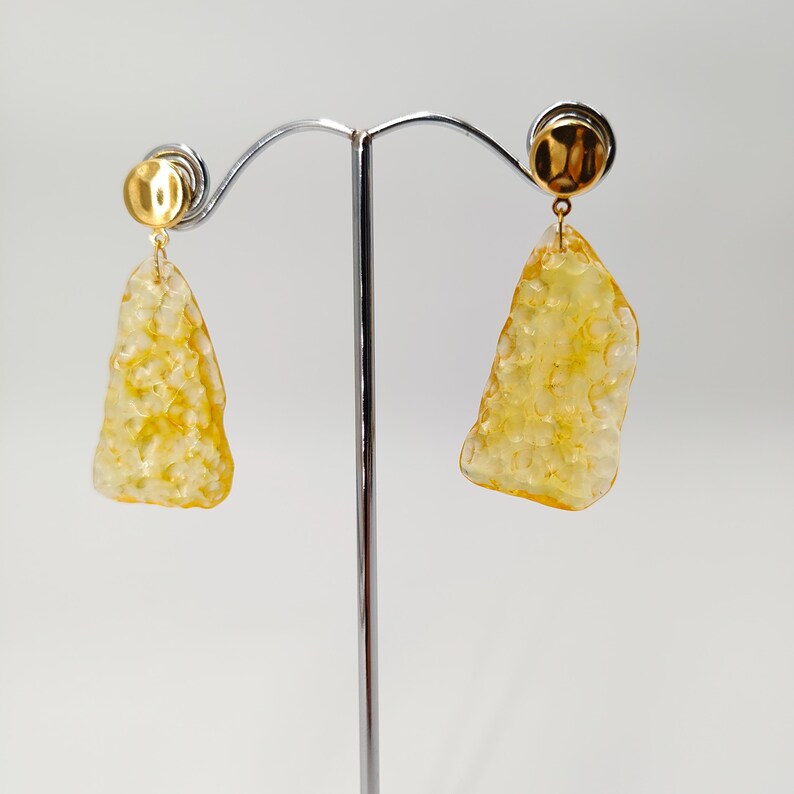 May include: A pair of gold-colored earrings with a textured, irregular, yellow-orange pendant. The earrings are displayed on a silver stand against a white background. The earrings have a post-back closure.