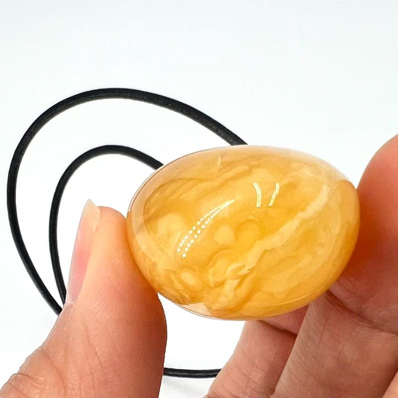 May include: A translucent, oval-shaped, amber-colored stone pendant with a black cord. The stone has a marbled appearance with lighter yellow and white swirls. The pendant is held by a person's hand against a white background.