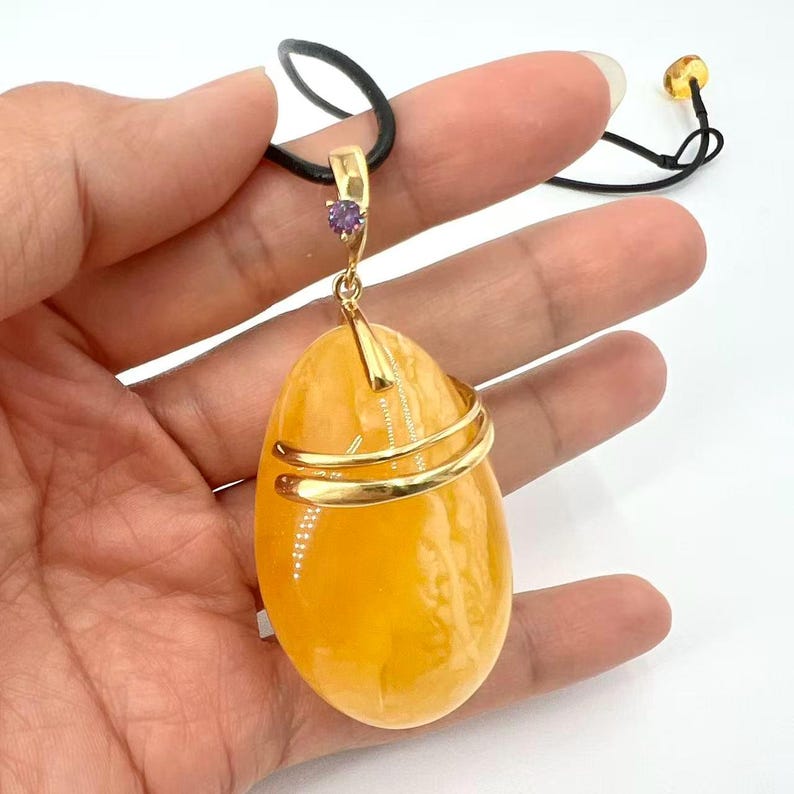 May include: A large, oval amber pendant with a gold-colored metal wrap and a small purple gemstone. The pendant hangs from a black cord necklace. The amber is a translucent, warm yellow-orange color.