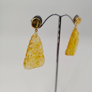 May include: A pair of gold-colored earrings with a textured, amber-colored, triangular pendant. The earrings are displayed on a silver earring stand against a white background. The earrings have a gold-colored stud with a circular design.