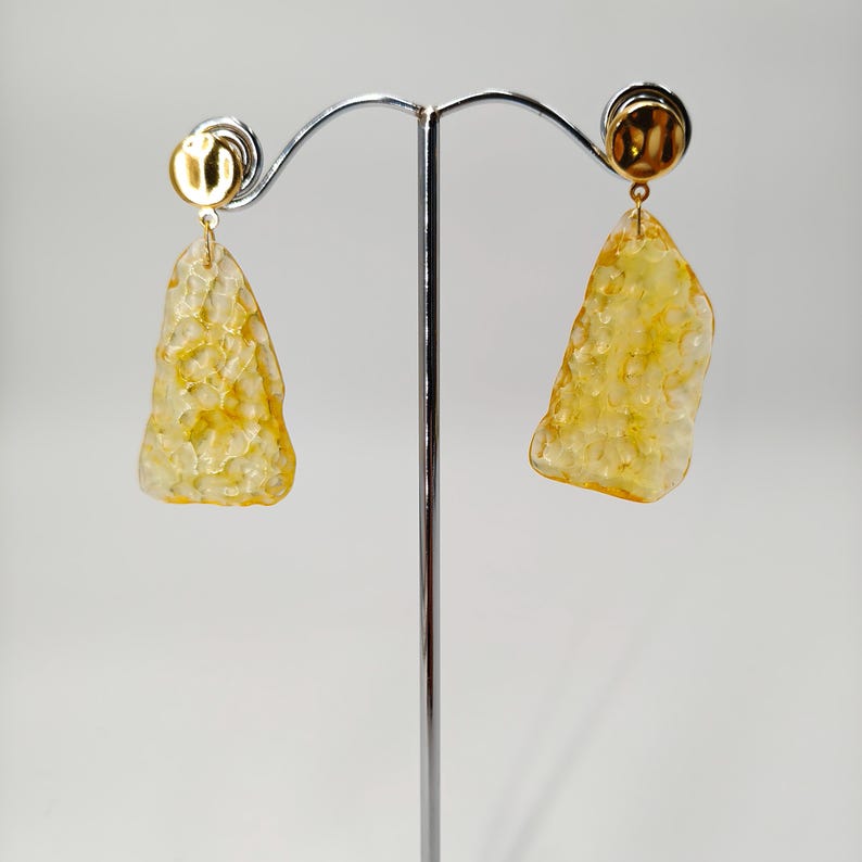 May include: A pair of gold-toned earrings with a textured, amber-colored, irregular-shaped pendant. The earrings are displayed on a silver jewelry stand against a white background. The earrings have a post-style backing.