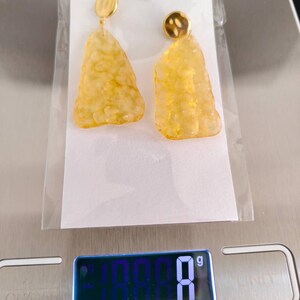 May include: A pair of gold-toned earrings with a textured, amber-colored, irregular shape. The earrings are displayed on a white card inside a clear plastic bag. The digital scale reads 8 grams.
