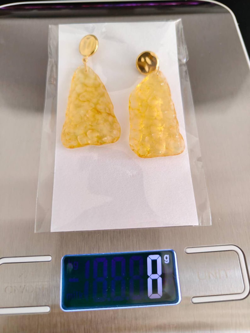 May include: A pair of gold-toned earrings with a textured, amber-colored, irregular shape. The earrings are displayed on a white card inside a clear plastic bag. The digital scale reads 8 grams.