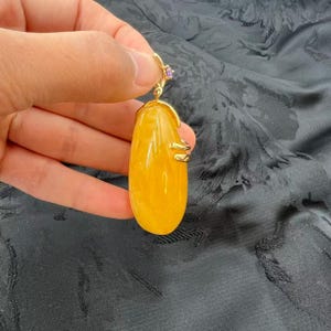 May include: A gold-toned pendant featuring a large, oval-shaped, amber-colored stone. The pendant has a gold bail with a small purple gemstone accent. The stone is approximately 2 inches long.