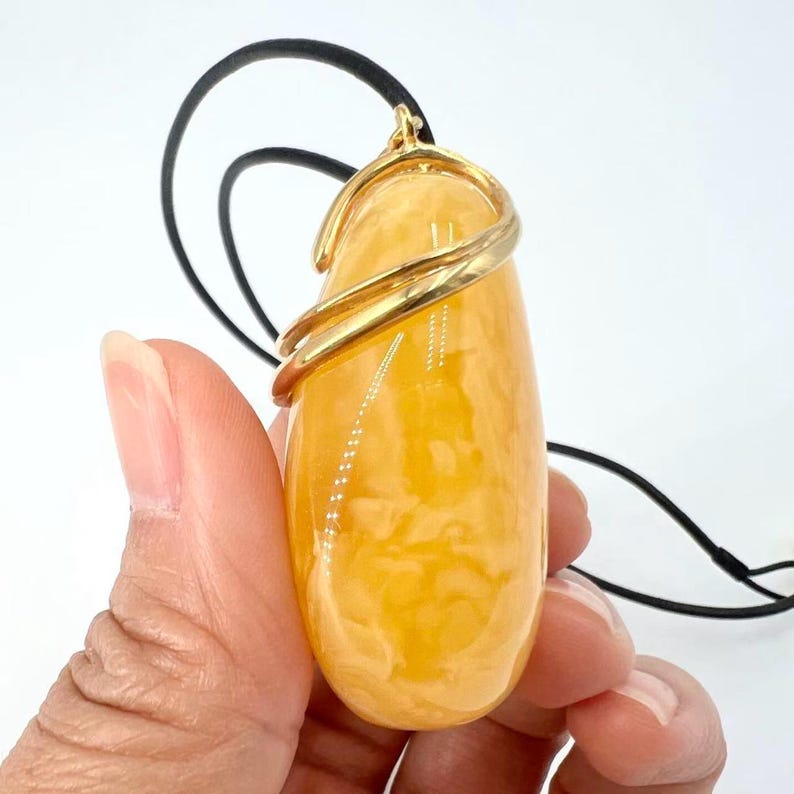 May include: A golden-yellow pendant with a teardrop shape, wrapped with a gold-colored metal band. The pendant hangs from a black cord. The background is white.