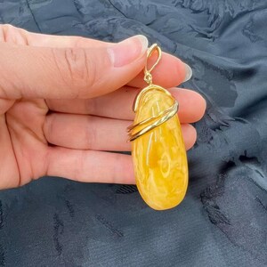 May include: A teardrop-shaped amber pendant with a gold-colored bail and wire wrapping. The amber is a translucent yellow with white marbling. The pendant is suspended from a gold-colored clasp.