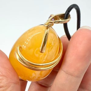 May include: A gold pendant with a large, oval, amber-colored stone. The pendant has a gold bail with a small, purple gemstone and is suspended from a black cord. The stone is encased in a gold setting.