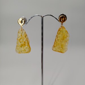 May include: A pair of gold-toned earrings with a textured, amber-colored, irregular shape. The earrings are suspended from a small, round gold stud. The earrings are displayed on a silver earring stand.