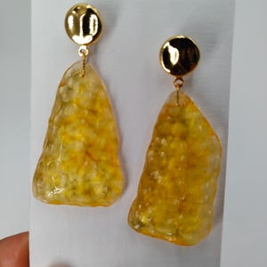 May include: A pair of gold-toned earrings. Each earring features a small, round stud and a textured, yellow, irregular-shaped pendant. The pendants have a translucent quality, with visible internal patterns. The earrings are displayed against a white background.