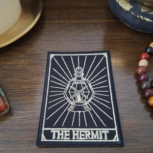 Può includere: Toppa ricamata in bianco e nero raffigurante la carta dei tarocchi dell'Eremita. Il disegno include una lanterna con linee radianti e la scritta "THE HERMIT". La toppa è rettangolare.